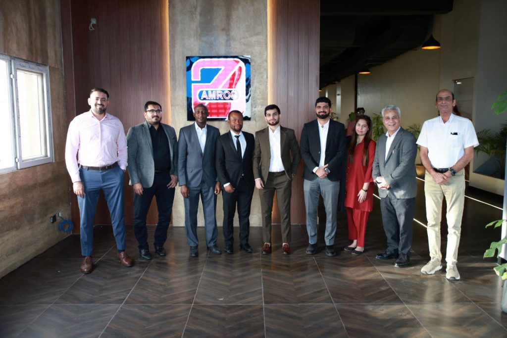 A group of professionals posing together in the lobby of Amros Pharmaceuticals during the Nigeria Inspection 2025 visit, standing in front of the company logo in a modern corporate setting.